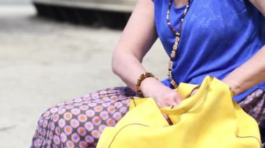 Yaşlı bir kadın, şık giyinmiş, parkta bir bankta sarı deri çantasında bir şey arıyor ve oradan bir telefon çıkarıyor, insanlar teknoloji kullanıyor, yüksek kalite 4K görüntü.