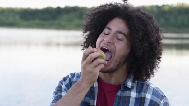 快乐的黑头发卷曲的英俊迷人的阿拉伯人在远足的过程中吃着青苹果 开心的笑着 看着相机 在湖面的背景下 高质量的Fullhd镜头 — 图库视频影像