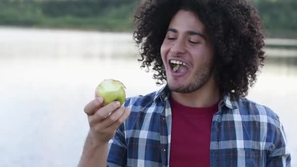 幸せな黒髪のカーリーミレニアルアラブの男はリンゴを噛んで白い健康な歯を披露し 喜びと高品質のフルHd映像でカメラを見る — ストック動画