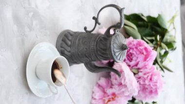 Kadınların eli, porselen bardaktaki espresso kahveyi gri çaydanlık ve pembe şakayıklı yeşil çaydanlığın arka planıyla karıştırıyor.