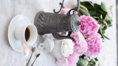 Kadınların eli espresso kahveyi karıştırıyor ve porselen fincana gümüş cımbızla şeker ekliyor. Masada pembe şakayıklar, yüksek kaliteli 4K görüntüler var.