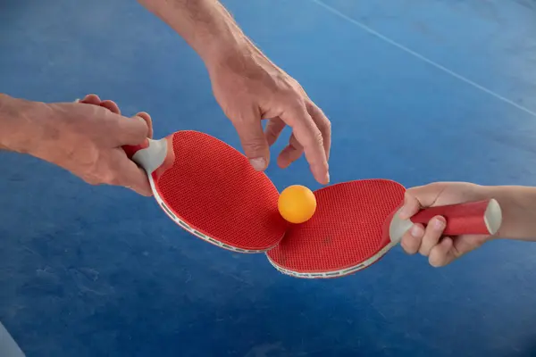 stock image male and childs hands hold red ping pong rackets with a yellow ball on a tennis table outdoors in sunny weather, training to play table tennis