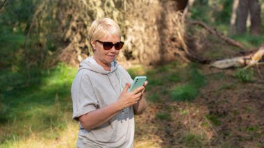 Ormanda bej örgü elbiseli bir kadın günlük adımlarını atıyor, çam ormanında havayı soluyor, akıllı telefonundaki yol haritasına bakıyor ve adım sayacını başlatıyor.