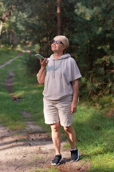 stock image woman in the forest in beige knitted suit takes her daily steps, breathes the air of a pine forest, records an audio message on a smartphone, blogs on social networks, 5G internet