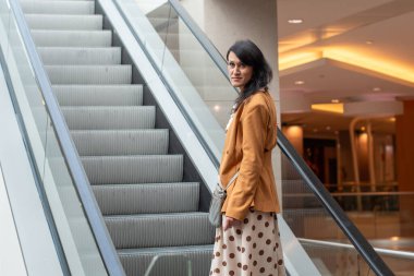 Kendinden emin genç esmer, alışveriş merkezinin üst katındaki yürüyen merdivenden yukarı çıkar, kameraya bakar, arka planı bulanıktır, yüksek kaliteli fotoğraf...