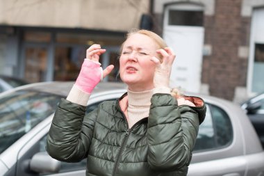 Yeşil ceketli üzgün bir kadın ve sağ kolunda pembe fiberglas bir bandajla duygusal çöküntüyle otoparkta ağlıyor, ellerini havaya kaldırıyor, araba süremiyor, kendini sınırlı hissediyor.