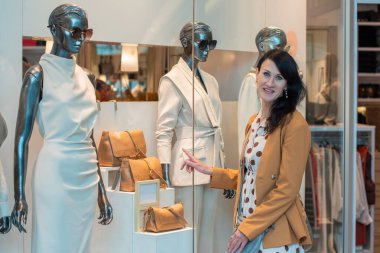Kendine güvenen genç esmer, lüks çantalar ve şık kıyafetler giymiş mankenlerle dolu bir vitrine bakar. Modern alışveriş merkezindeki bir butikte, yüksek kaliteli bir fotoğrafla.