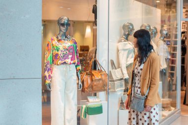 Kendine güvenen genç esmer, lüks çantalar ve şık kıyafetler giymiş mankenlerle dolu bir vitrine bakar. Modern alışveriş merkezindeki bir butikte, yüksek kaliteli bir fotoğrafla.