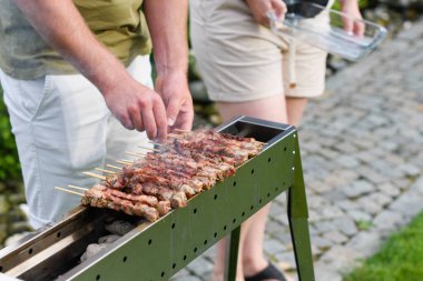 young man grilling meat kebabs on coals, ARROSTICINI - typical southern Italian cuisine, a luxurious picnic on the terrace near the house in summer  clipart