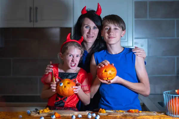 stock image Mother, daughter and son in Halloween costumes carve eyes and teeth on pumpkins in a darkened room, traditional autumn decorations for the holiday