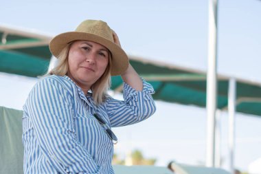 Genç bir kadın kumsalda güneşli bir yerde oturuyor. Şapka takıyor, gergin görünüyor. Tatilde bile dinlenemiyor. 