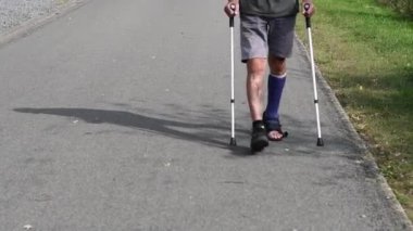 Modern mavi fiberglas alçılı sol bacağı kırık bir adam yürüyüş ayakkabısıyla yürüyor, iki bacağı üzerinde yürüyor, koltuk değneklerine yaslanıyor yaralı bacağındaki yükü alıyor, rehabilitasyon konseptini değiştiriyor.,