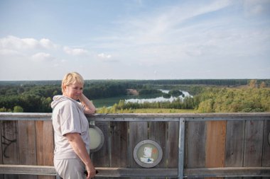 middle aged blonde with short haircut poses in front of view from observation tower of beautiful lake in Lommel-Sahara Nature Park in Belgium, sunny autumn day clipart