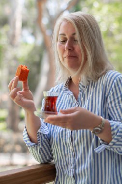 Sarışın, beyaz bir kadın balkonda Türk Armudu bardağından çay içiyor ve portakallı pastadan bir ısırık alıyor. Öğle yemeğinden sonra lezzetli bir tatlı. Yaz tatilinde geleneksel oryantal tatlı.