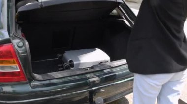 Genç esmer kadın, güneşli bir günde parlak bir arabanın bagajından parlak bir gümüş bavul çıkarır ve yaz tatili boyunca seyahat etmeyi dört gözle bekler.,