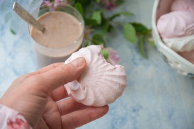 Isırılmış vanilya şekerlemesi tutan kadın eli, bir vazo dolusu vanilyalı marshmallow ve bir bardak sütlü kahve, masanın üzerinde pembe çiçek dalları olan vazonun yanında,