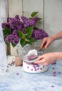 Kadın eli kesme keki, leylak çiçekleriyle süslenmiş vazonun yanında, ahşap masa üzerinde leylak dalları olan, çiçekli ve kırsal yaşam tarzını vurgulayan, narin, durgun bir hayat.,