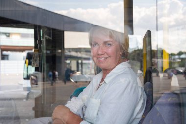 Otobüste oturan orta yaşlı beyaz kadın, otobüs durağı ve toplu taşıma araçlarının yansıması ve odak dışı insanlar.,