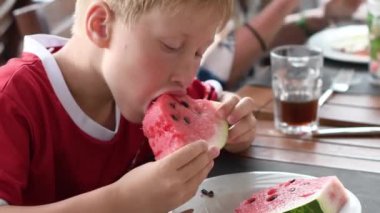 Kırmızı tişörtlü sarışın çocuk, yaz tatili sırasında bir kafede bir parça karpuz yiyor, soğuk sulu meyvelerle susuzluğunu gideriyor.,