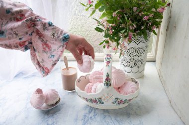 Kadın eli vanilya şekerlemesi tutuyor, narin, vazo dolusu vanilyalı marşmelov ve bir bardak sütlü kahve, vazonun yanında ahşap masa üzerinde pembe çiçek dalları, üst manzara