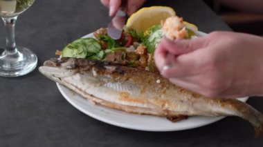 Tanınmayan bir adam pilav, taze salata ve limon dilimleri ile servis edilen kızarmış levrek yiyor. Masada bir bardak beyaz şarap var.,