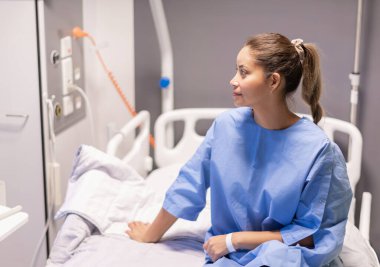 Hastane koğuşunda mavi elbiseler giymiş, doktor tarafından muayene edilmeyi bekleyen, servis ve hizmetlerden nazikçe gülümseyen genç bir kadın hasta.