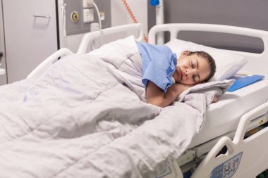 Mavi bornozlu kliniğin genç bayan hastası hastane yatağında uyuyor. Sakin bir ortamda, özel odada. Hafif ışıklandırma, iyileşme ve bakım kavramı.,