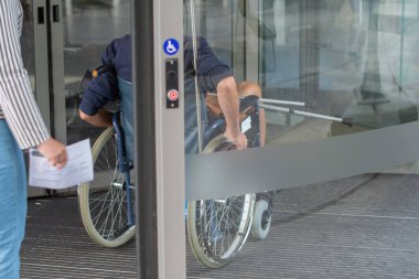 Tekerlekli sandalyede oturan orta yaşlı bir adam kliniğin camlı kapısına doğru ilerliyor. Ellerini tekerlekleri kontrol etmek için kullanıyor. Karısı da onu takip ediyor.,