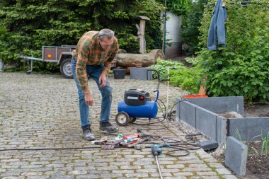 Orta yaşlı bir adam bahçesinde sera inşa etmek için çeşitli aletler kullanıyor, ev yapımı sebze yetiştirme konsepti, yeşil bitki zeminine karşı kaldırımlı bir taş yolda duruyor.