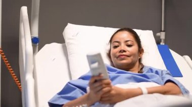 Mavi elbiseli genç hasta, yastık yüksekliğini uzaktan kumandalı işlevsel elektrikli yatakta ayarlıyor, rahat bir dinlenme yeri bulmak için kaldırıyor ve indiriyor, kadın gülümsüyor ve eğleniyor.,