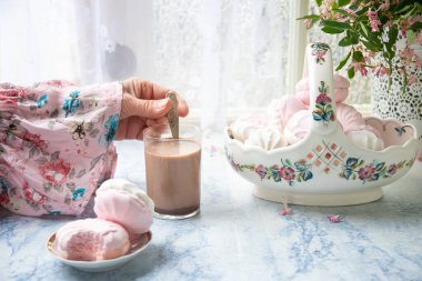 Kadın eli, masada sütle kahve karıştırıyor vazonun yanında da bir sepet vanilyalı marşmelov ve pembe çiçeklerle, köy hayatı kavramı.,