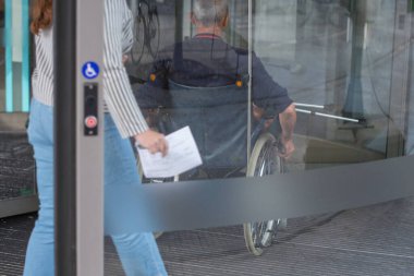 Tekerlekli sandalyede oturan orta yaşlı bir adam kliniğin camlı kapısına doğru ilerliyor. Ellerini tekerlekleri kontrol etmek için kullanıyor. Karısı da onu takip ediyor.,