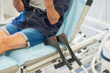 Adam, klinikteki tıbbi kanepede derin bir düşünceye dalmış doktorun onu muayene etmesini ve kırık bacağını alçıya almasını bekliyor. Koltuk değnekleri yatağın yanında yatıyor.,