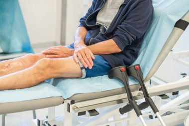 Adam klinikte bir tıp yatağında oturuyor, günlük giysiler giyiyor, koltuk değnekleri yanında, sağlık sigortası ayarında.,