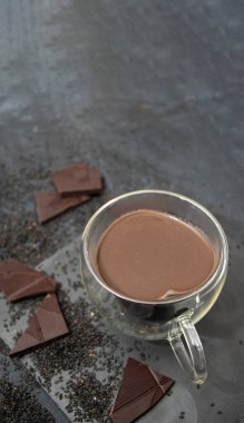 Çikolata parçaları ve gri dokulu arka planda siyah chia tohumları ile çevrili masada kalp şeklinde cam bardak sıcak çikolata, 2025 yılı çöl konsepti, moka mus, kopyalama alanı 