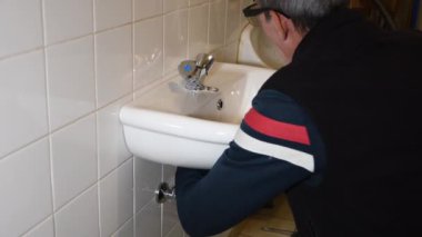 Gözlüklü bir adam, banyodaki fayanslı duvarda yeni beyaz seramik bir lavaboyla dikkatlice asılıyor ve tamir ediliyor. Ev işi kıyafetleri içinde, su tesisatı konseptinde çalışıyor.,