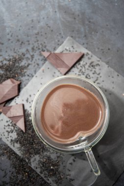 Çikolata parçaları ve gri dokulu arka planda siyah chia tohumları ile çevrili masada kalp şeklinde cam bardak sıcak çikolata, 2025 yılı çöl konsepti, moka mus, kopyalama alanı 