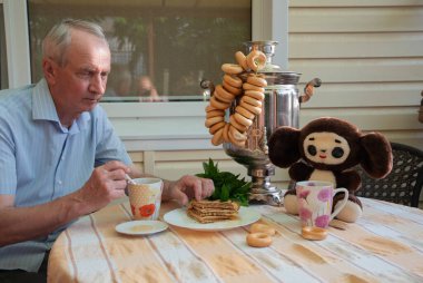 an elderly man enjoys a traditional Russian tea party with a samovar, pretzels, pancakes with honey in the company of a plush toy Cheburashka at the table,  clipart