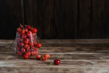 Olgun kırmızı kirazlar taşar küçük bir cam kavanoz ve metal saplı bir tahta masa üzerinde koyu ahşap bir zemin üzerinde oturarak basit ama çekici bir hayat yaratır.,