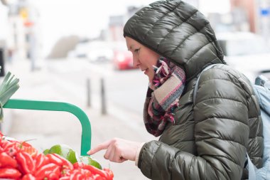 Sağ kolunda pembe fiberglas olan kadın bulutlu bir günde marketten taze sebze seçiyor, kapüşonlu ceket giyiyor, kısıtlamalara rağmen aktif bir hayat sürüyor.