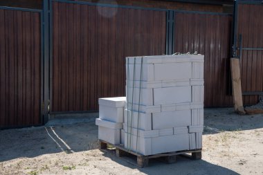 İnşaat alanındaki ahşap paletlere bağlanmış hafif havalandırmalı beton bloklar inşaata başlamaya hazır, arka planda güneşli bir günde ahşap tahıl metal çitler var.,