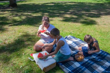 Cheerful family enjoys refreshing summer picnic outdoors,mother eats sweet watermelon slices,older son cuts fruit,younger one eats chips on blue plaid blanket while playing game on smartphone,