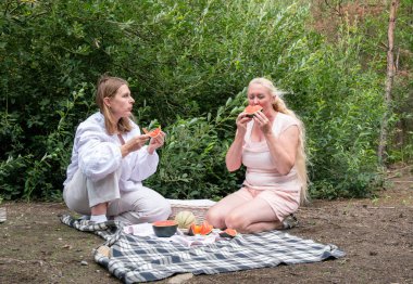 Two adult women enjoying a relaxing summer picnic in nature, sitting on a plaid blanket, eating refreshing slices of watermelon and melon against a backdrop of green foliage
