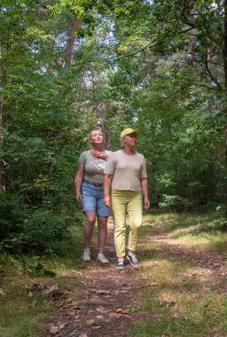 Two older women walking on a dirt path in a lush green forest, one woman placing a hand on the others shoulder, both looking upwards, sharing a moment of calm and companionship outdoors