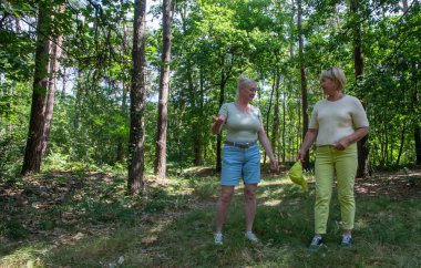 Two mature female friends are engaged in conversation and smiling while enjoying a leisurely walk in a vibrant green forest, finding joy and connection in nature during their outdoor activity