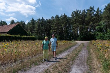 Senior couple enjoying their retirement by walking through a field of drying sunflowers on a dirt path, with a forest and a red roofed house in the background