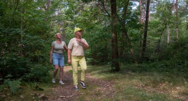 Two senior women enjoying a healthy and active lifestyle, walking along a rustic path in a beautiful green forest, engaging in outdoor leisure activities and spending quality time together