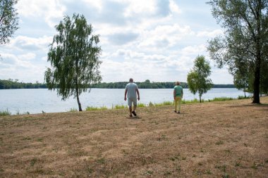 Mature couple strolling along dry grass by a tranquil lake at midday, enjoying summer leisure, peaceful companionship and open sky on a relaxing outdoor walk