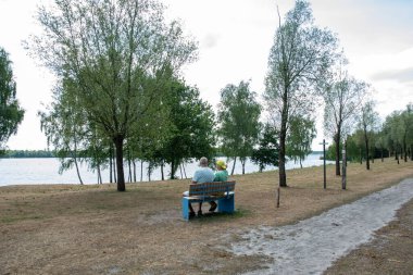 Senior couple sitting on a blue park bench overlooking a calm lake, surrounded by green trees and dry grass, spending quality time together in a peaceful natural environment during daylight