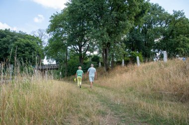 Senior couple walking together on a dry grassy path in a rural landscape during summer, finding leisure and enjoying the outdoors amidst tall grass and green trees, with a bridge in the background
